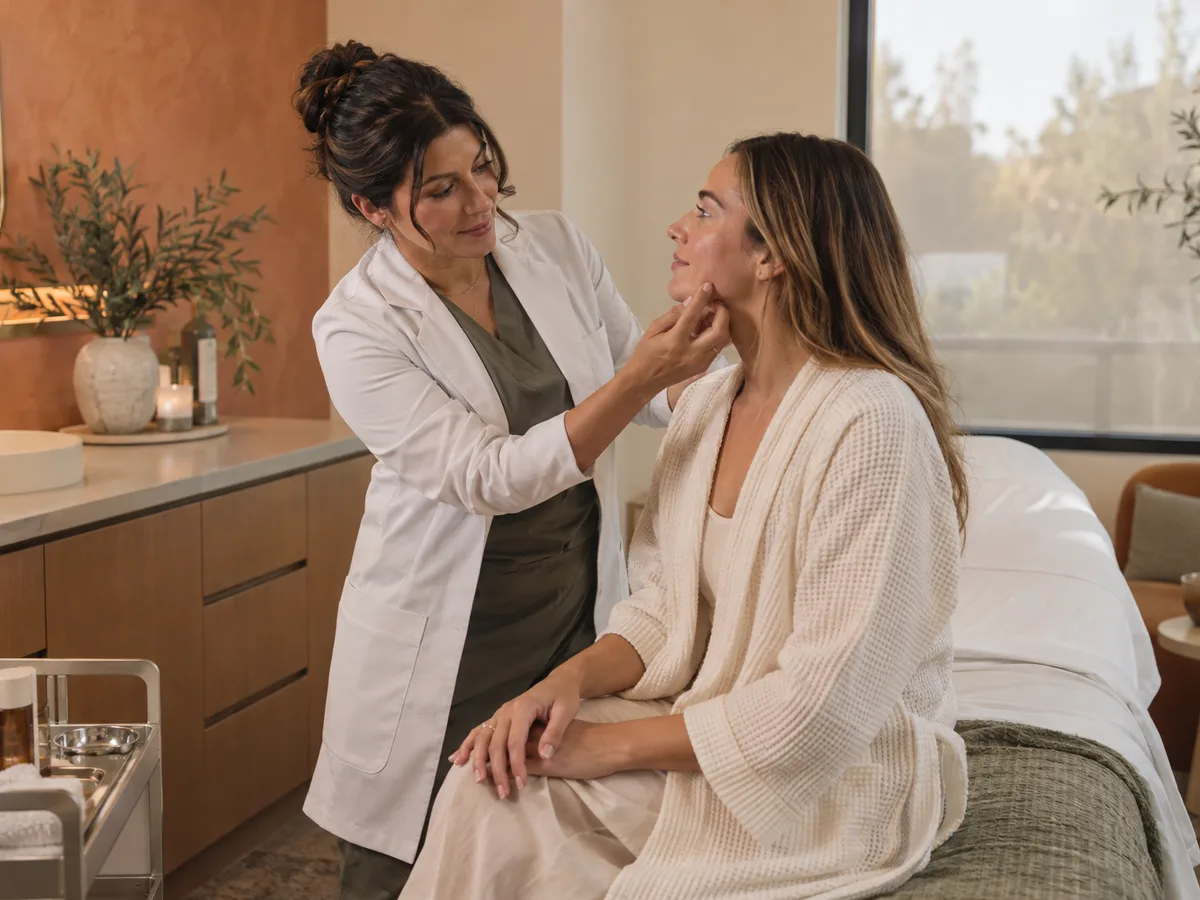 A woman in a white coat holds the jaw of a woman seated on a bed